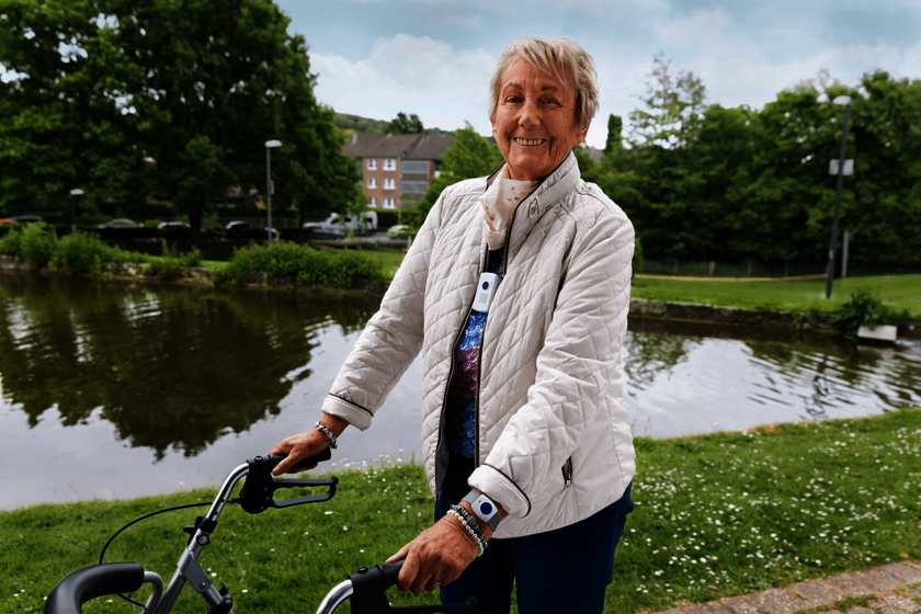 Lächelnde ältere Dame in weißer Steppjacke und Rollator in einem Parkt, im Hintergrund ein See. Um den Hals und am Handgelenk trägt sie einen Notrufknopf