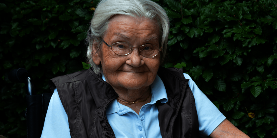 eine ältere Dame in hellblauem Shirt und grauer Weste sitzt im Garten und lächelt in die Kamera