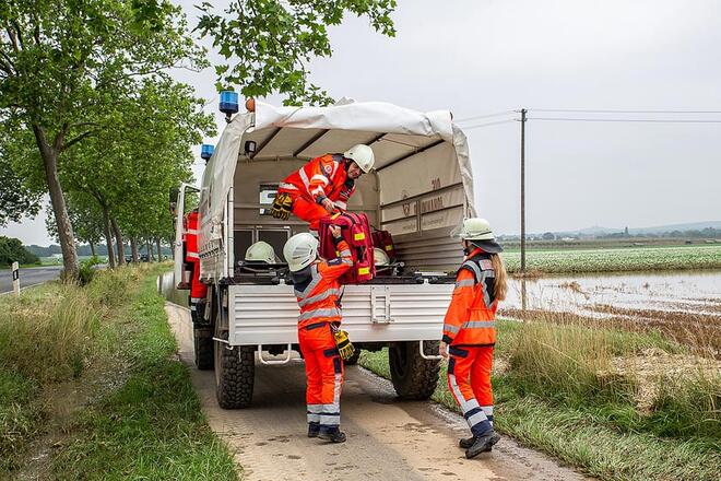 3 Einsatzkräfte stehen neben einem Einsatzfahrzeug auf einer Landstraße und laden Sachen aus.