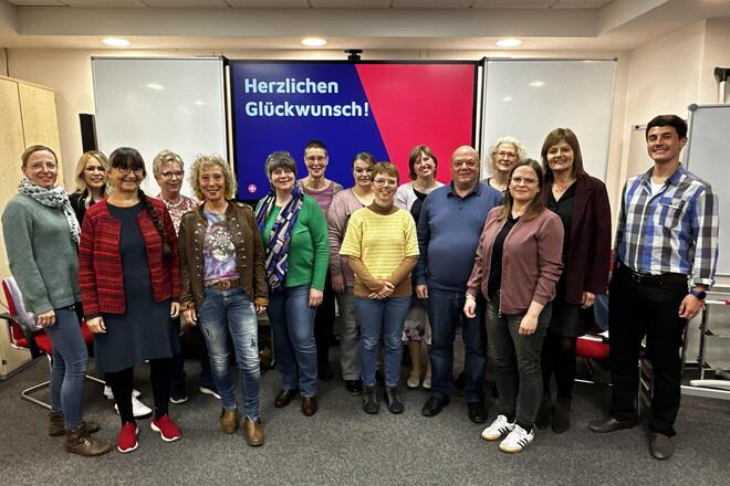 Gruppenfoto mit 15 Personen: 13 der insgesamt 14 Teilnehmenden des 27. Vorbereitungskurses mit Leitender Koordinatorin Julia Kunz (links) sowie Koordinatorin Felicitas Zimmermann (Mitte)