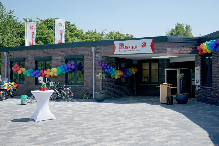 Vorplatz und Eingangsbereichs eines einstöckigen Gebäudes mit dunklen Klinkern und dunkelgrauen Fensterrahmen geschmückt mit Johanniter-Fahnen und Luftballons