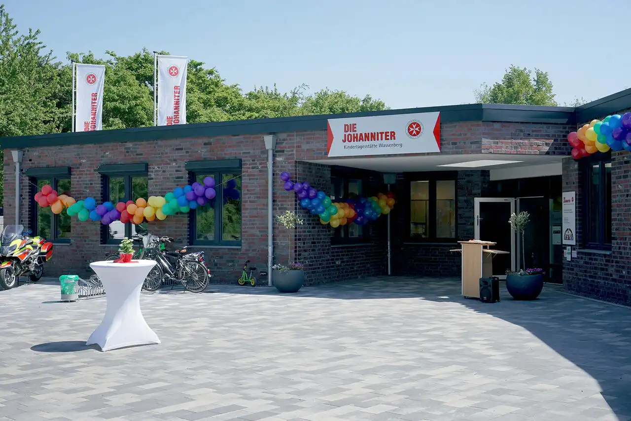 Vorplatz und Eingangsbereichs eines einstöckigen Gebäudes mit dunklen Klinkern und dunkelgrauen Fensterrahmen geschmückt mit Johanniter-Fahnen und Luftballons