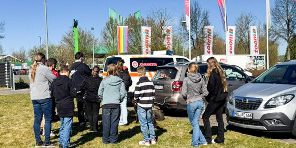 Kinder stehen auf einer Wiese und hören einer Frau zu