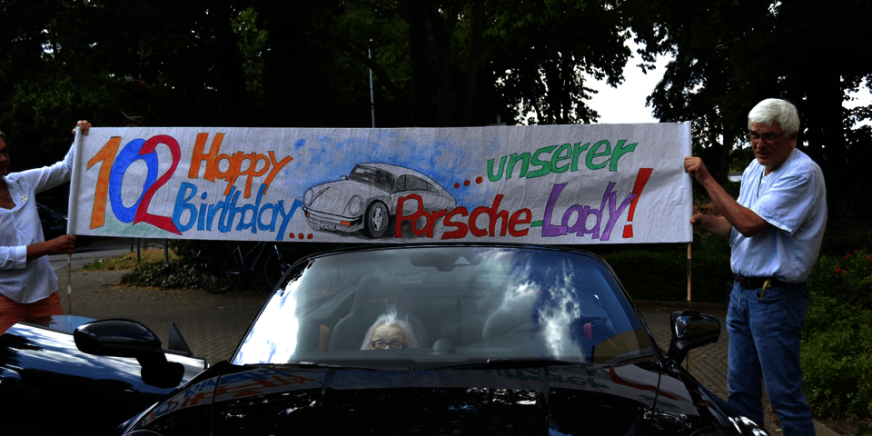 Eine Frau und ein Mann halten über das Dach eines Porsches ein Banner mit der Aufschrift "102 Happy Birthday unserer Porsche-Lady"