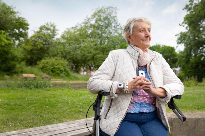 Eine Seniorin sitzt in einem Park auf einem Rollator. Um den Hals sowie am Handgelenk trägt sie ein rechteckiges weißes Gerät mit einem blauen Knopf