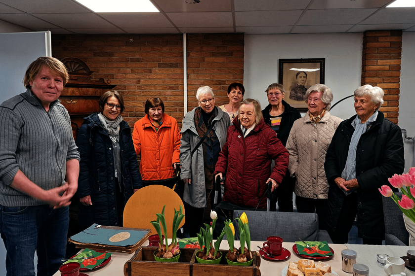Neun Frauen und ein Mann stehen an einem mit Blumen geschmückten Kaffeetisch. Auf einer Ecke liegt eine Kladde mit alten Papieren. 