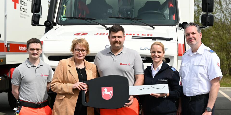 Zwei junge Männer, eine Frau und ein weiterer Mann in Johanniter-Kleidung. Eine Frau im beigen Blazer hält mit einem Mann einen großen Pappschlüssel. Alle stehen vor einem Fahrzeug