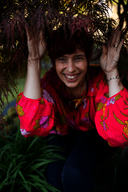 eine Frau mit rotem Haar hält Äste über dem Kopf und lächelt im Garten