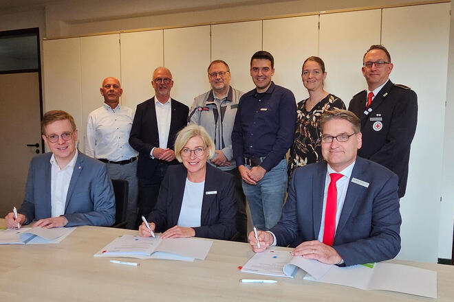 Gruppe Menschen sitzen bzw. stehen um einen Tisch herum. Drei von ihnen unterschreiben einen Vertrag.