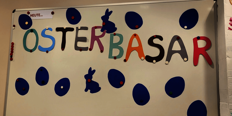 Auf einer weißen Tafel steht in bunten Buchstaben "Osterbasar". Darum blaue Osterhasen und Eier