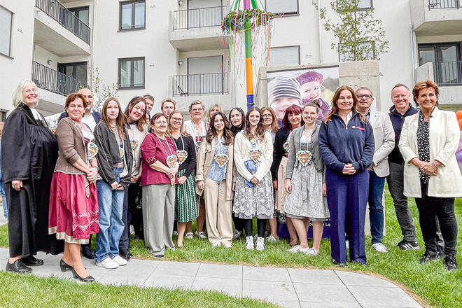 Mehrere Leute stehen vor einem bunten Maibaum, einige haben ein Lebkuchenherz um.