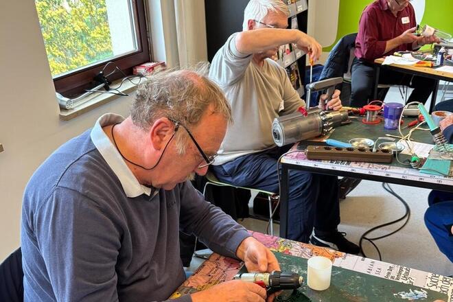 Drei ältere Männer reparieren gemeinsam Elektrogeräte an Tischen im Johanniter-Repaircafé. Sie nutzen Werkzeug und konzentrieren sich auf die Reparatur verschiedener Alltagsgegenstände in einem Gemeinschaftsraum.