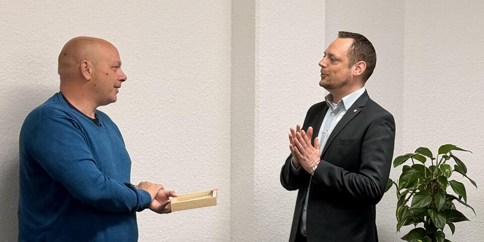 Ein Mann hält ein Geschenk in der Hand. Ein anderer spricht mit ihm. 