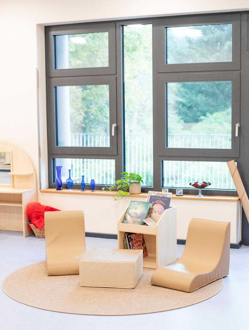 Zwei Sitzliegen mit Hocker und Bücherregal vor einem großen Fenster.