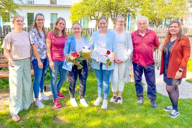 Mehrere Leute stehen in einem grünen, sonnigen Garten. Zwei von ihnen halten ein Geschenk und Blumen.