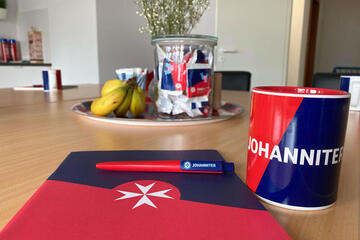 Ein Block mit Kugelschreiber und daneben einer Kaffeetasse in den Farben rot/blau der Johanniter laden zum verweilen oder arbeiten im Begegnungsraum Liederbach ein.