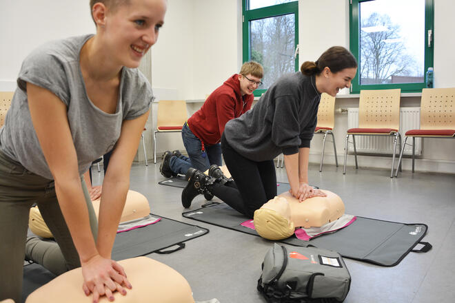 Drei junge Menschen üben die Reanimation an Übungspuppen und lachen dabei.