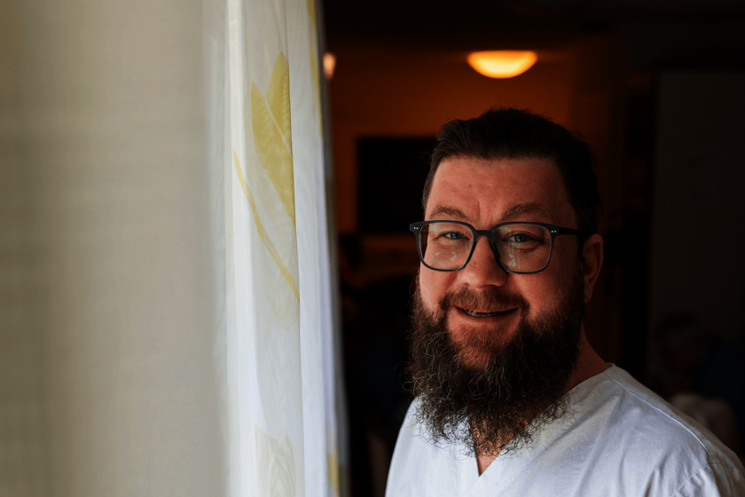 Ein Mann in weißer Arbeitskleidung steht am Fenster neben einem Vorhang, lächelt 