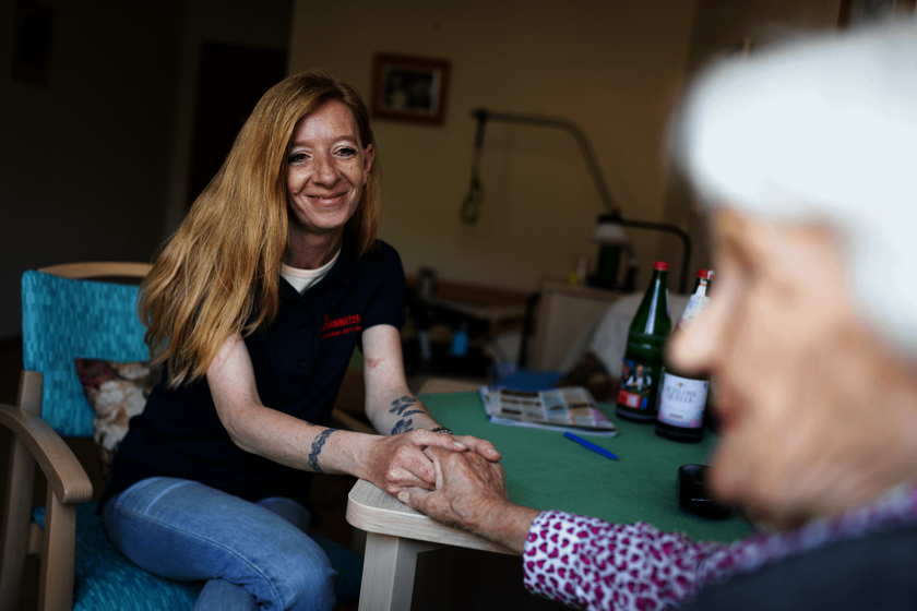 Pflegerin hält Hand einer älteren Frau am Tisch, freundliche Atmosphäre im Pflegeheim.
