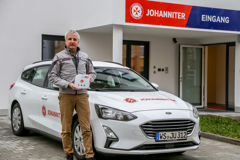 Johanniter-Mitarbeiter steht mit Hausnotrufgerät vor Dienstwagen und Eingang des Johanniter-Zentrums in Wasserburg.
