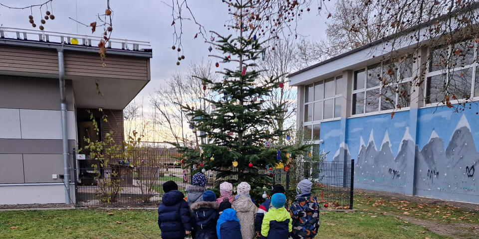 Kinder vor einem geschmückten Weihnachtsbaum draußen. Weihnachtstrucker-Pakete stehen daneben