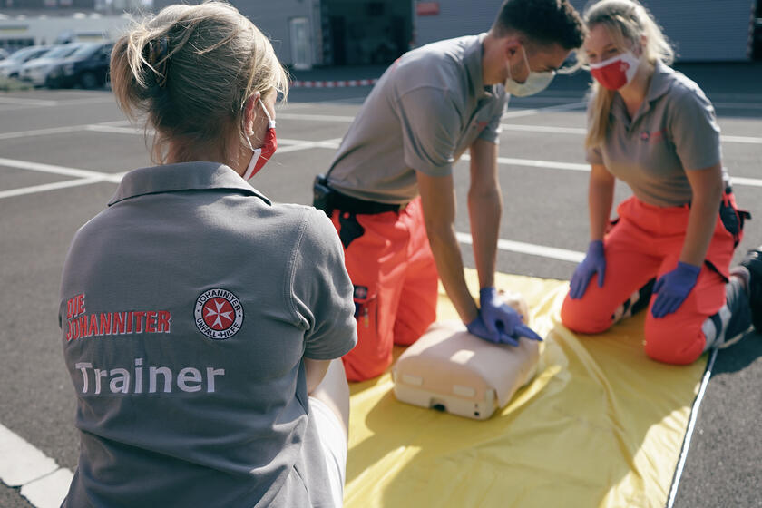 Eine Johanniter-Trainerin in grauem Poloshirt beobachtet zwei Personen, die an einer Puppe Wiederbelebung trainieren