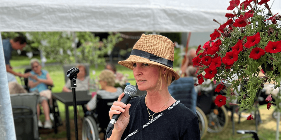 Ein junge Frau mit Strohhut hält in einem Garten ein Mikrofon; im Hintergrund Gartentische