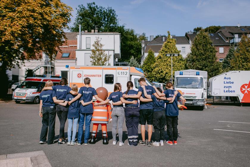 Mitglieder der Johanniter-Jugend stehen gemeinsam, Arm in Arm vor einem Rettungswagen. Man sieht ihre Rücken.