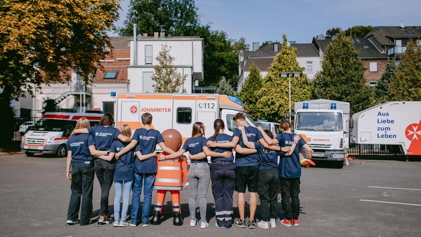 Mitglieder der Johanniter-Jugend stehen gemeinsam, Arm in Arm vor einem Rettungswagen. Man sieht ihre Rücken.