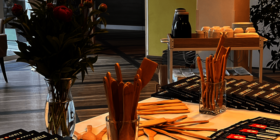 auf einem Tisch liegen Bücher mit der Aufschrift "Leib und Seele". Daneben sind Kochlöffel aus Holz und eine Vase mit Pfingstrosen.