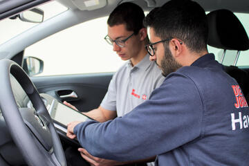 Zwei Männer im Auto; einer zeigt dem anderen Informationen auf einem Tablet.