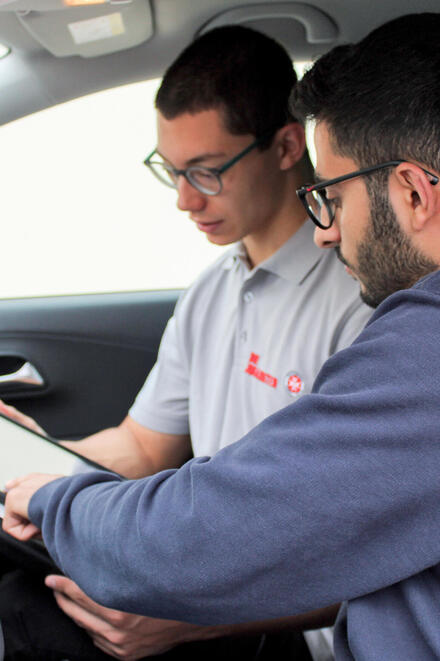 Zwei Männer im Auto; einer zeigt dem anderen Informationen auf einem Tablet.