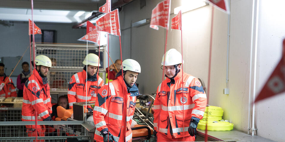 Vier Johanniter in Einsatzkleidung tragen eine Rettungstrage durch einen Hindernisparcours.