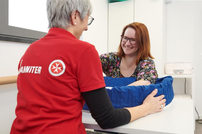 Eine Mitarbeiterin der Johanniter in rotem Poloshirt mit Logo hilft einer lächelnden Frau, die an einem Tisch sitzt. Die Frau hat rotbraune Haare und trägt eine Brille sowie ein buntes, geblümtes Oberteil. Ihre Hände befinden sich in einer Fühlkiste. Die Szene findet in einem hellen Raum statt.