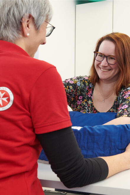 Eine Mitarbeiterin der Johanniter in rotem Poloshirt mit Logo hilft einer lächelnden Frau, die an einem Tisch sitzt. Die Frau hat rotbraune Haare und trägt eine Brille sowie ein buntes, geblümtes Oberteil. Ihre Hände befinden sich in einer Fühlkiste. Die Szene findet in einem hellen Raum statt.