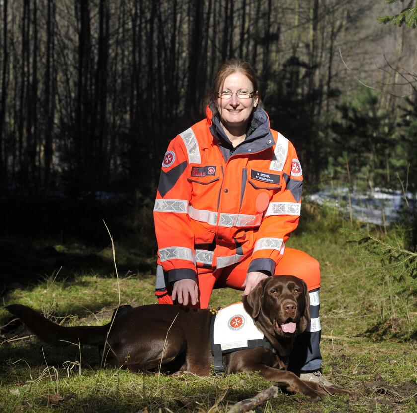 Eine Rettungshundeführende der Johanniter kniet in leuchtender Einsatzkleidung im Wald neben ihrem braunen Labrador. Der Hund trägt eine Einsatzweste und liegt entspannt im Gras. Beide schauen freundlich in die Kamera.