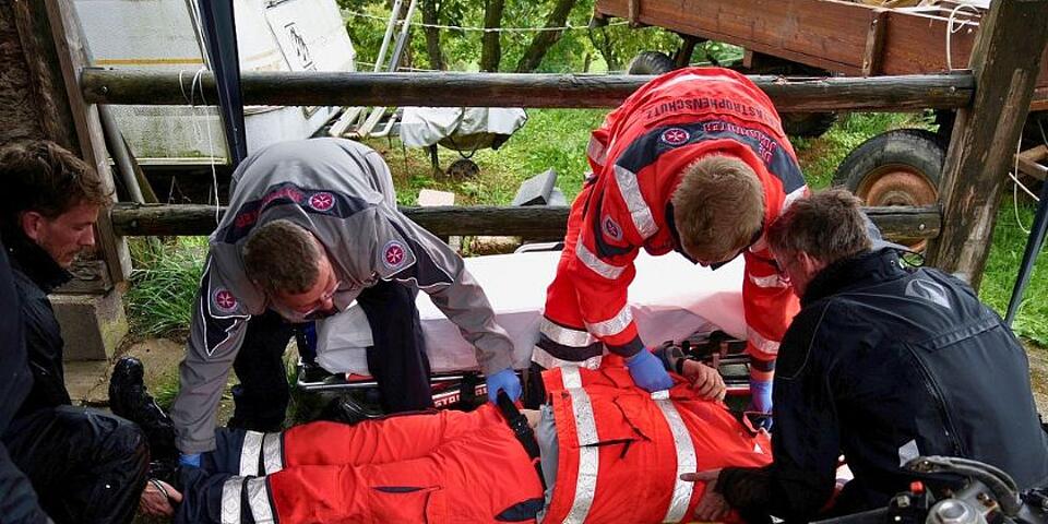 Das Unfallopfer liegt auf einem Spineboard. Offenbar wurde er gerade darauf gehoben: Zwei Motorradfahrer packen noch an Kopf- und Fußende an, zwei Johanniter greifen an Gürtel, Bein und Schulter.