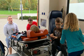 Ein Jugendgruppenleiter fährt eine Trage in einen Rettungswagen, während Kinder zuschauen