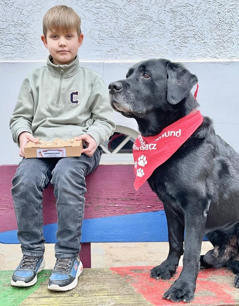 Ein Junge sitzt vor einer Wand und neben ihm ein schwarzer Lesehund mit Halstuch.