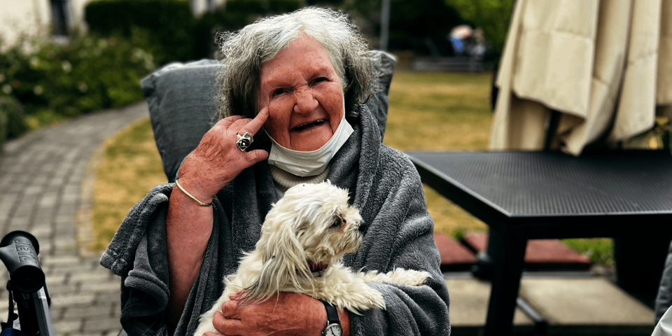 Ein Frau mit weißen Haaren ist in eine graue Decke gehüllt und hält einen weißen kleinen Hund auf dem Schoß.