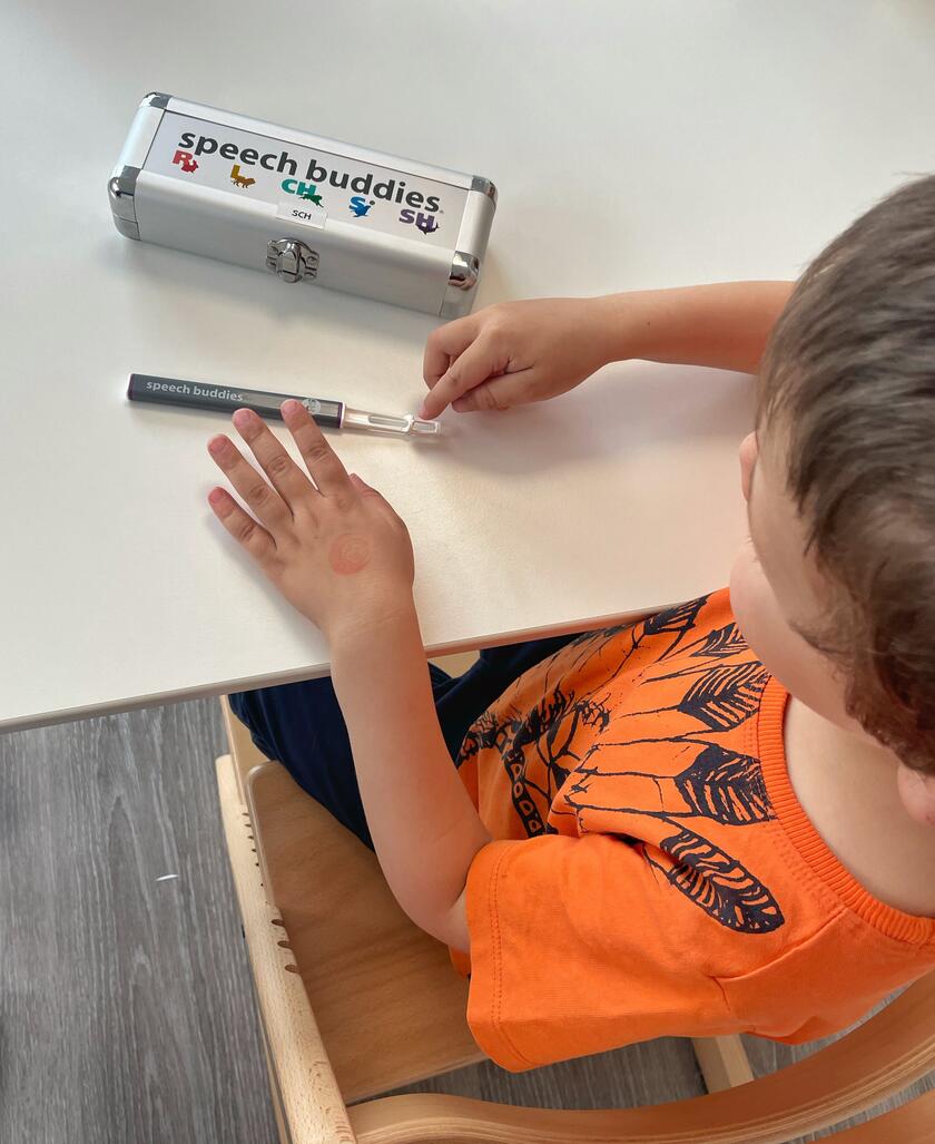 Ein Kind mit orangefarbenem T-Shirt sitzt an einem Tisch und hält ein Logopädie-Hilfsmittel namens „speech buddies“ in der Hand. Auf dem Tisch liegt ein silbernes Etui mit der Aufschrift „speech buddies“ und bunten Symbolen.