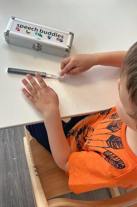 Ein Kind mit orangefarbenem T-Shirt sitzt an einem Tisch und hält ein Logopädie-Hilfsmittel namens „speech buddies“ in der Hand. Auf dem Tisch liegt ein silbernes Etui mit der Aufschrift „speech buddies“ und bunten Symbolen.