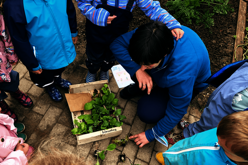Kinder und eine hockende Frau mit dunklen Haaren beschäftigen sich mit einer Holzkiste mit kleinen Pflanzen, die zwischen ihnen auf dem Boden steht.