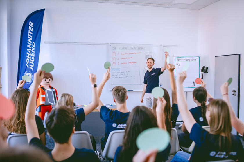 Jugendliche auf Stühlen vor einer Tafel, Flippchart und einem Referenten melden sich.