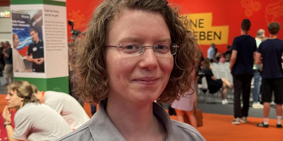 Eine Frau mit Brille und lockigem Haar in einer Messehalle. Sie trägt ein Johanniter-Poloshirt