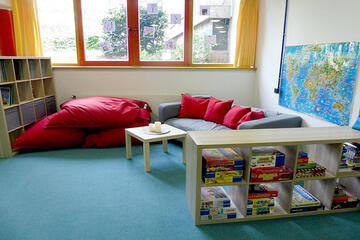 Sitzgelegenheit mit gemütlichen großen roten Kissen. Im Vordergrund ist ein Regal mit zahlreichen Gesellschaftsspielen zu sehen.