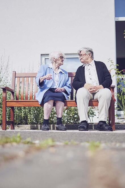 Eine Bewohnerin und ei Bewohner sitzen auf einer Parkbank