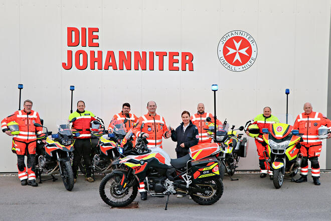 Mehrere Johanniter stehen vor einer Wand mit einem großen Johanniter Logo und haben Motorräder um sich stehen.