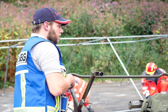 Ein Johanniter steht vor einem Metall Gestell. Im Hintergrund sieht man weitere Johanniter in Einsatzkleidung.