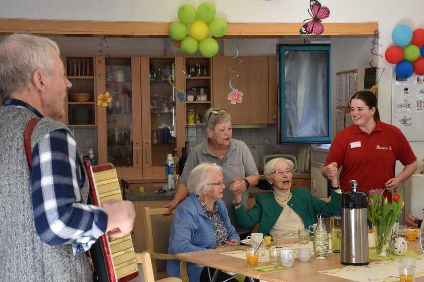 zwei Seniorinnen feiern das Frühlingsfest, hinter ihnen stehen zwei Pflegekräfte, im Vordergrund spielt ein Akkordeonspieler